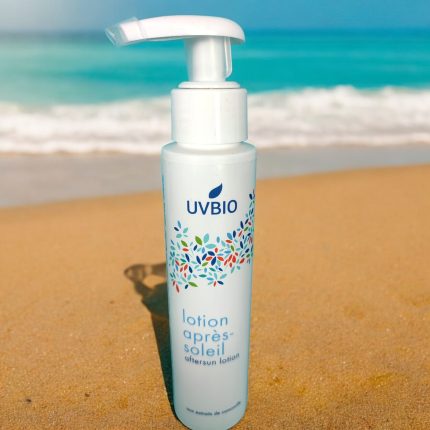 Bouteille de lotion après-soleil UVBIO sur la plage, avec eau et sable, sans nanoparticules dans l’image.