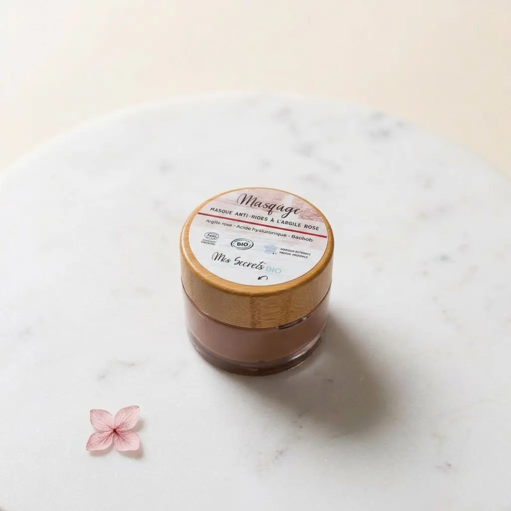 Masque anti-âge à l’argile rose « Masq’âge » 50 ml dans son pot, posé sur table marbrée avec petite fleur rose à côté.